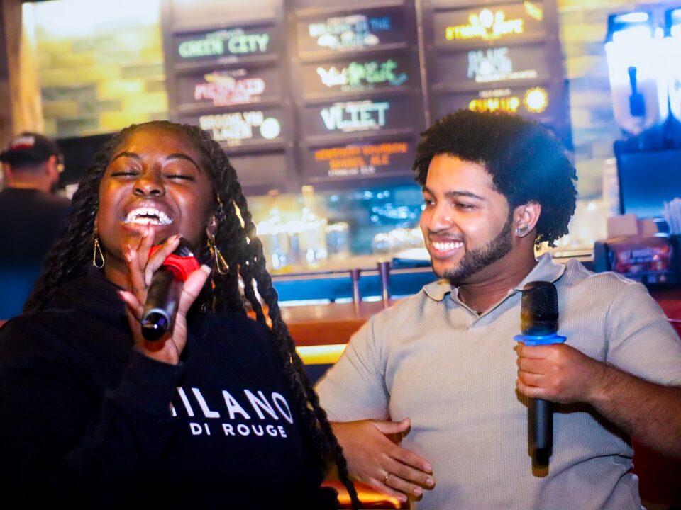 Two friends at a bar laughing and singing into handheld microphones, woman in a black hoodie on the left and a smiling man in a light gray shirt on the right, with neon menu boards behind them.