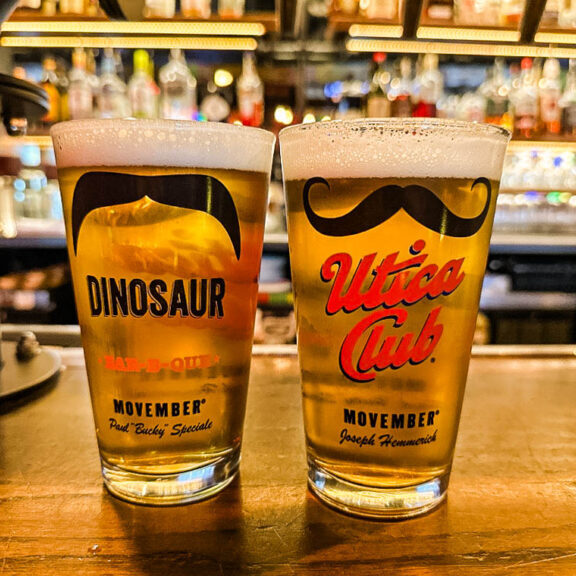 Two pint glasses filled with beer sit on a wooden bar counter, each featuring a black mustache graphic on the top. The left glass has 'DINOSAUR' in bold yellow letters with 'Movember' and 'Paul "Bucky" Speciale' written below. The right glass has 'Utica Club' in red cursive font with 'Movember' and 'Joseph Hemmerick' written beneath. The background showcases a well-stocked bar with liquor bottles and beer taps, softly blurred.